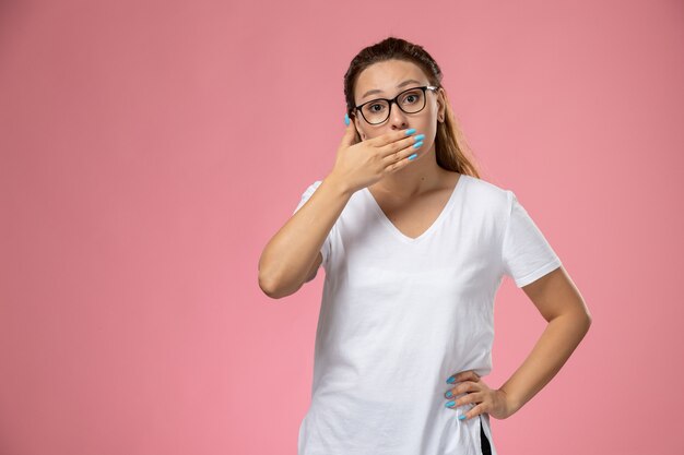 Giovane femmina attraente di vista frontale in maglietta bianca che posa appena con la bocca coperta sui precedenti rosa