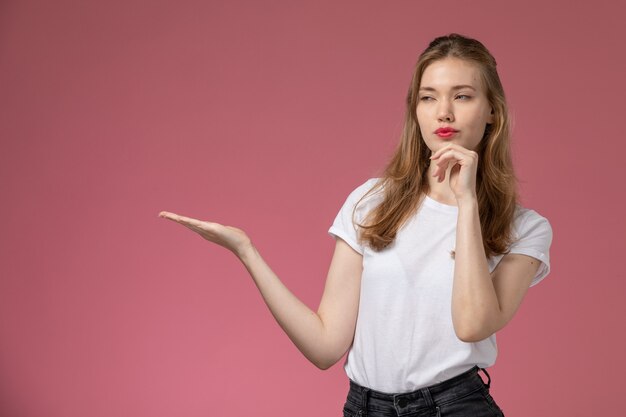 Giovane femmina attraente di vista frontale in maglietta bianca che posa con l'espressione di pensiero sulla foto a colori di posa femminile del modello della parete rosa