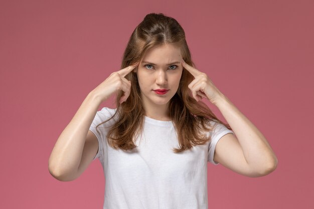 Giovane femmina attraente di vista frontale in maglietta bianca che posa con la posa di pensiero sulla foto a colori di posa femminile del modello della parete rosa