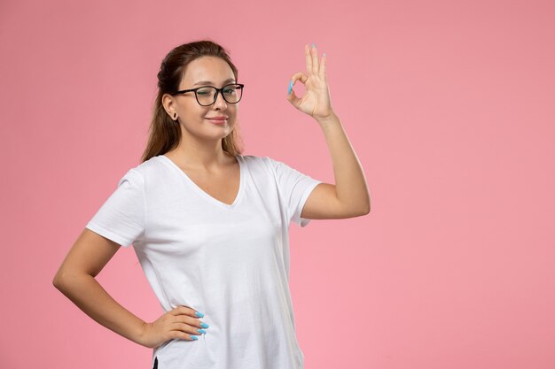 Giovane femmina attraente di vista frontale in maglietta bianca che posa con un leggero sorriso e ammiccante sullo sfondo rosa