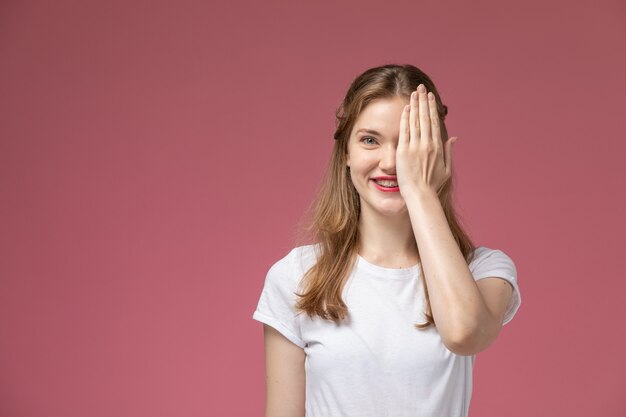 Giovane femmina attraente di vista frontale in maglietta bianca che sorride e che copre metà del suo viso sul muro rosa modello di colore femminile giovane