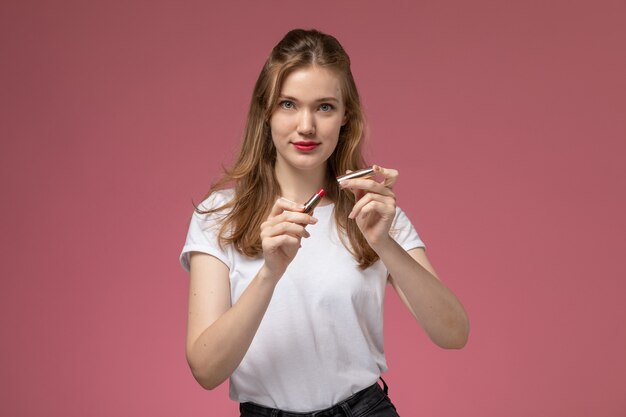 Giovane femmina attraente di vista frontale in maglietta bianca che tiene il rossetto sulla ragazza della femmina di colore del modello della parete rosa scuro