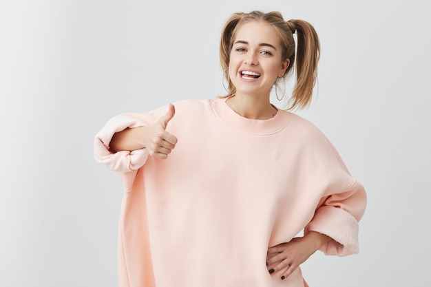 Giovane femmina caucasica emozionante allegra con due code di cavallo, in maglione rosa, mostrando gesto giusto e sorridere, dimostrando i suoi denti bianchi, godendo della sua vita spensierata. Va tutto bene!