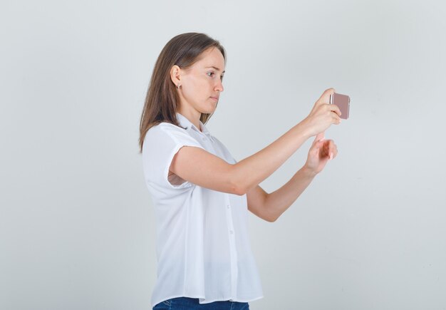 Giovane femmina che cattura foto su smartphone in camicia bianca, jeans e sguardo concentrato.