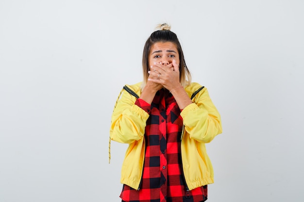 Giovane femmina che copre la bocca con le mani in camicia a scacchi, giacca e sembra scioccata. vista frontale.