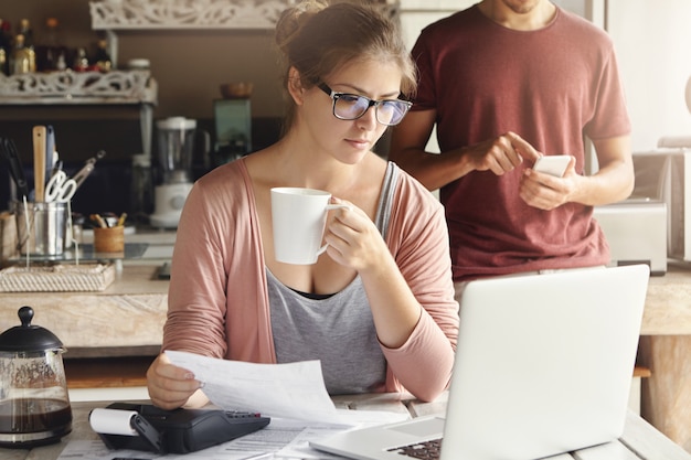 Giovane femmina che ha espressione concentrata guardando lo schermo del portatile aperto, tenendo la carta e la tazza di caffè in mano durante il calcolo delle spese domestiche