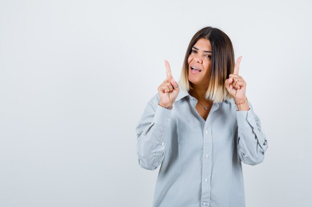 Giovane femmina che indica in su in camicia surdimensionata e che sembra felice, vista frontale.