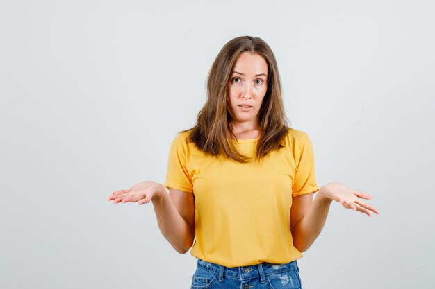 Giovane femmina che mostra gesto impotente in maglietta, pantaloncini e sguardo confuso. vista frontale.