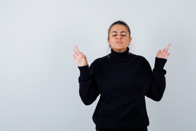 Giovane femmina che mostra il gesto di meditazione in maglione a collo alto e che sembra pacifica. vista frontale.