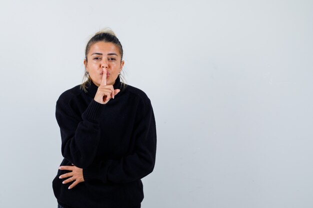 Giovane femmina che mostra il gesto di silenzio in maglione a collo alto nero e che sembra sicura. vista frontale.