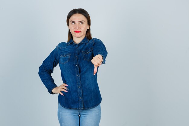 Giovane femmina che mostra il pollice verso il basso mantenendo la mano sul fianco in camicia di jeans e jeans e guardando dispiaciuto