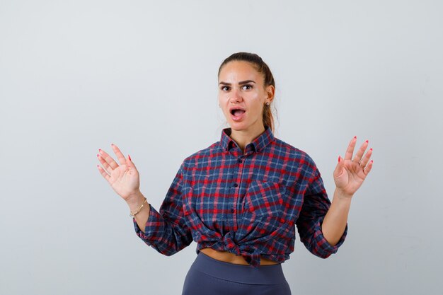 Giovane femmina che mostra le palme in gesto di resa in camicia a scacchi, pantaloni e sembra perplessa. vista frontale.