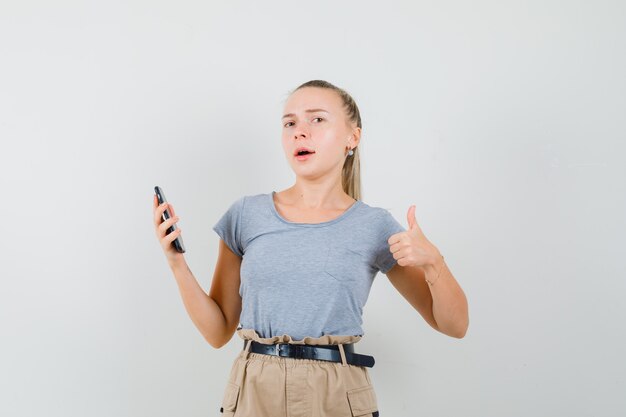 Giovane femmina che tiene il telefono cellulare, mostrando il pollice in su in t-shirt, pantaloni vista frontale.
