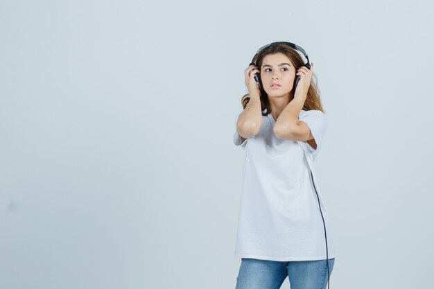 Giovane femmina che tiene le mani sulle sue cuffie in maglietta bianca, jeans e che sembra sognante, vista frontale.