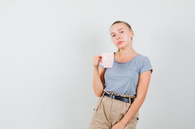 Giovane femmina che tiene tazza di bevanda in maglietta, pantaloni e un bell'aspetto. vista frontale.