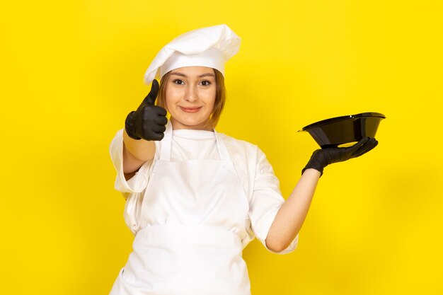 giovane femmina di cucina in abito bianco cuoco e berretto bianco in guanti neri mostrando ciotola nera