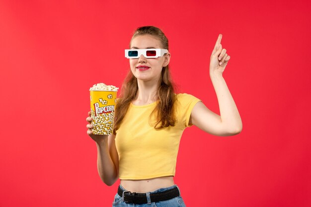 Giovane femmina di vista frontale al cinema che tiene popcorn in occhiali da sole d sul colore femminile del cinema del cinema del cinema della parete rossa