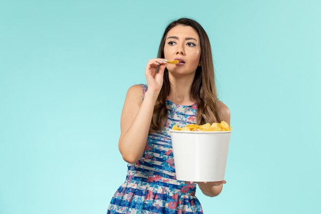 Giovane femmina di vista frontale che mangia cips e che guarda film sulla superficie blu