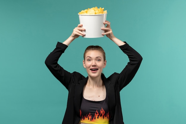 Giovane femmina di vista frontale che mangia le patatine fritte e che guarda film sulla superficie blu