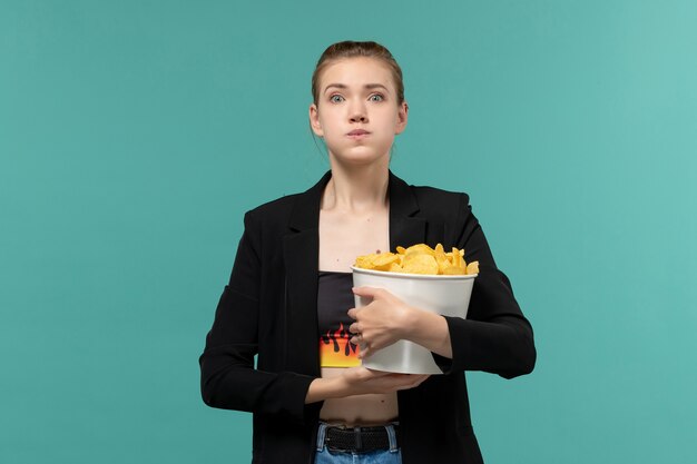 Giovane femmina di vista frontale che mangia patatine fritte che guarda film sullo scrittorio blu