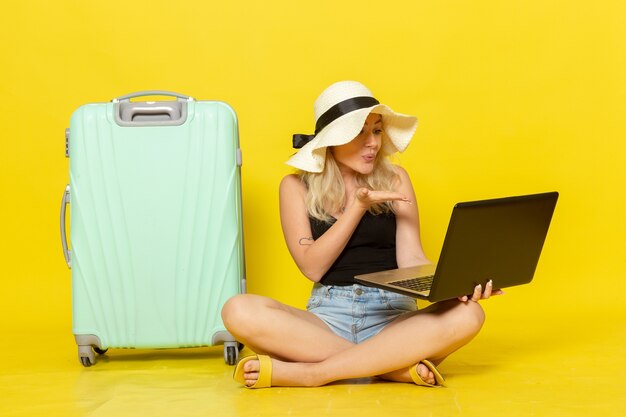 Giovane femmina di vista frontale che utilizza il suo computer portatile e che parla con qualcuno tramite video sul sole di viaggio di viaggio di viaggio di vacanza di viaggio della parete gialla