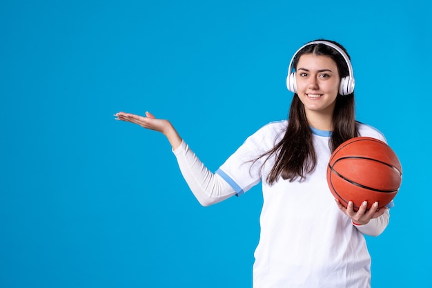 Giovane femmina di vista frontale con gli auricolari che tengono la pallacanestro sulla parete blu