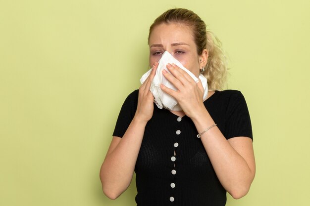 Giovane femmina di vista frontale con l'asciugamano bianco intorno alla sua gola che si sente molto malato e malato pulendosi il naso sulla salute femminile di malattia di malattia della parete verde chiaro