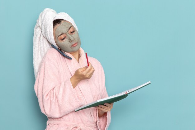 Giovane femmina di vista frontale in accappatoio rosa che parla sul telefono sulla doccia crema di selfcare di bellezza di pulizia della parete blu