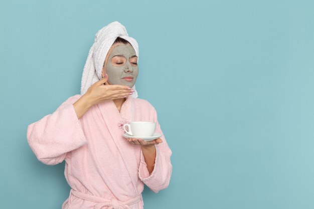 Giovane femmina di vista frontale in accappatoio rosa con la maschera sul suo fronte che tiene il caffè sulla crema di selfcare di bellezza di pulizia della doccia della parete blu