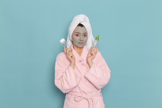 Giovane femmina di vista frontale in accappatoio rosa con la maschera sul suo viso che tiene gli spruzzi sulla crema di auto-cura di bellezza di pulizia della doccia della parete blu
