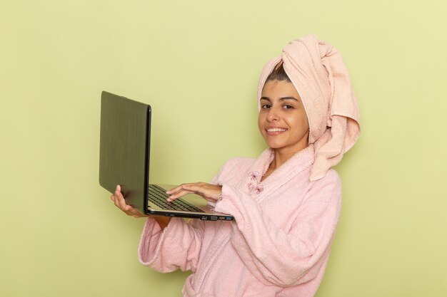 Giovane femmina di vista frontale in accappatoio rosa usando il computer portatile sulla superficie verde