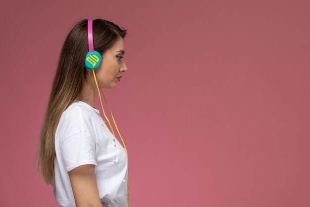 Giovane femmina di vista frontale in camicia bianca che ascolta la musica con i suoi auricolari sul muro rosa, donna di modello di posa della donna di colore