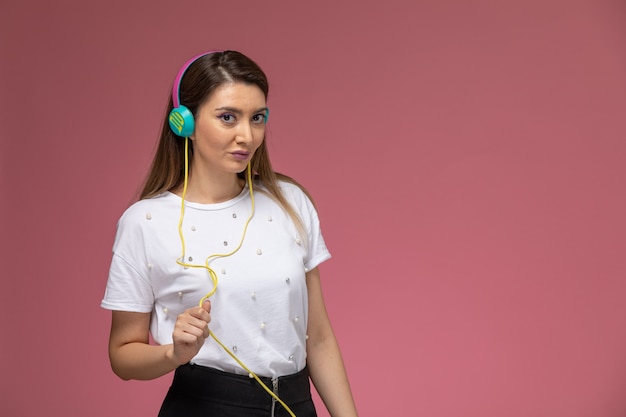 Giovane femmina di vista frontale in camicia bianca che ascolta la musica tramite auricolari colorati sulla parete rosa, modello di posa della donna di colore