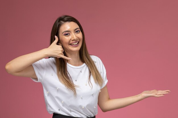 Giovane femmina di vista frontale in camicia bianca che mostra la posa di telefonata sulla parete rosa, modello di posa della donna di colore