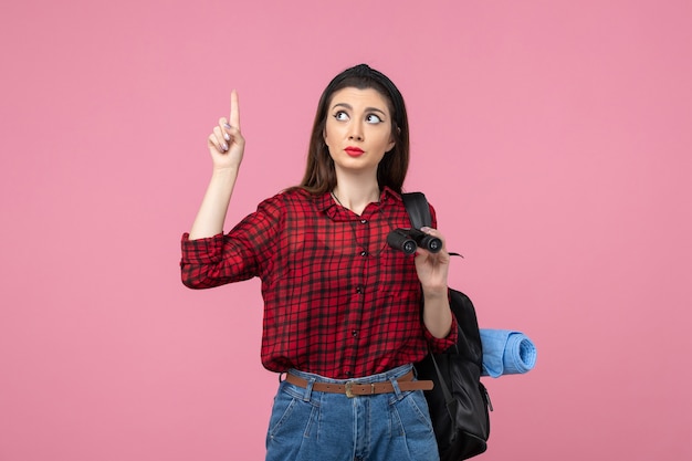 Giovane femmina di vista frontale in camicia rossa con il binocolo sullo studente di colore rosa della donna dello sfondo