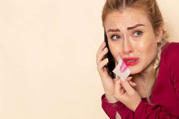 Giovane femmina di vista frontale in camicia rossa con la bocca legata che parla sul telefono sulla foto del panno femminile dello spazio crema