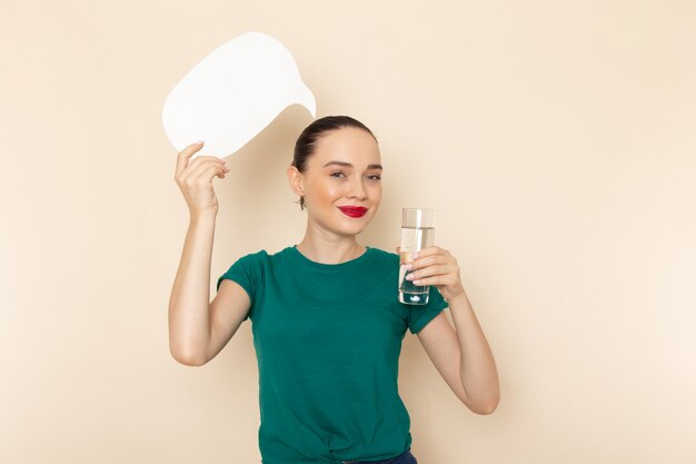 Giovane femmina di vista frontale in camicia verde scuro e blue jeans che tengono bicchiere d'acqua e segno bianco sul beige