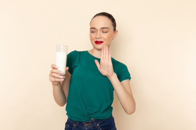 Giovane femmina di vista frontale in camicia verde scuro e blue jeans che tengono il bicchiere di latte che rifiuta di bere sul beige