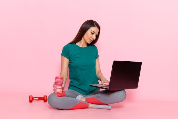 Giovane femmina di vista frontale in maglietta verde che lavora con il suo computer portatile sulla parete rosa chiaro vita sport esercizio allenamenti bellezza atleta femmina sottile