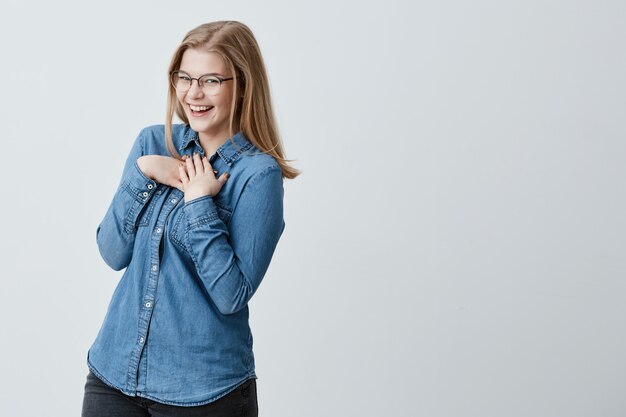 Giovane femmina europea civettuola giocosa affascinante con capelli lisci biondi che sorridono come se flirtando. Occhiali da portare della ragazza graziosa e camicia del denim che hanno espressione allegra sul suo fronte