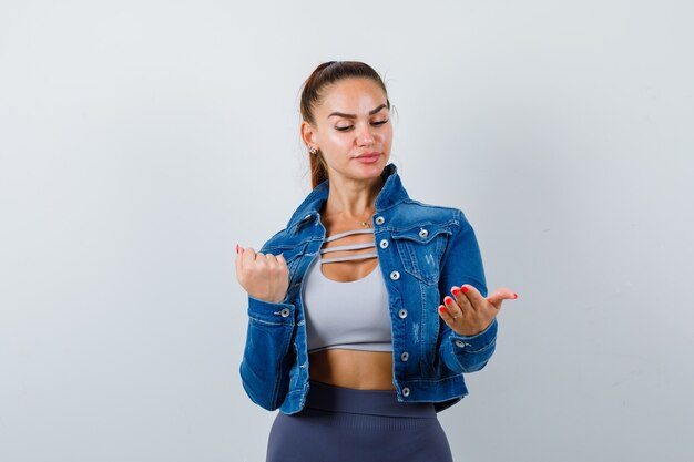 Giovane femmina in forma che guarda il palmo allargato mentre mostra il pugno chiuso in alto, giacca di jeans e sembra confuso, vista frontale.