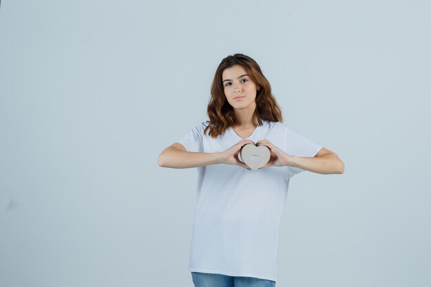 Giovane femmina in maglietta bianca, jeans che tengono il contenitore di regalo e che sembrano fiduciosi, vista frontale.