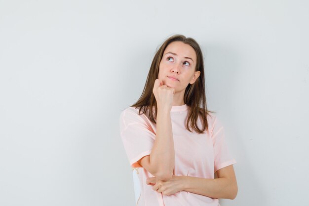 Giovane femmina puntellando il mento sul pugno in maglietta rosa e guardando pensieroso, vista frontale.