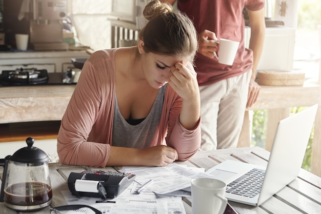 Giovane femmina sollecitata premurosa che si siede al tavolo da cucina con documenti e computer portatile che prova a lavorare attraverso la pila di fatture, frustrata dalla quantità di spese domestiche mentre facendo bilancio familiare