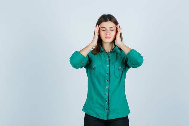 Giovane femmina strofinando le sue tempie in camicia verde e guardando rilassato. vista frontale.