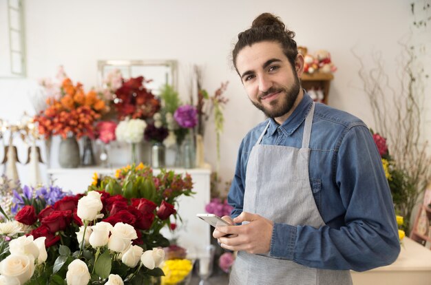 Giovane fiorista maschio felice che tiene telefono mobile a disposizione che guarda alla macchina fotografica