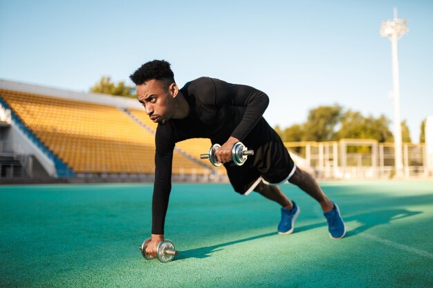 Giovane forte sportivo afroamericano che fa esercizi con manubri durante l'allenamento allo stadio cittadino