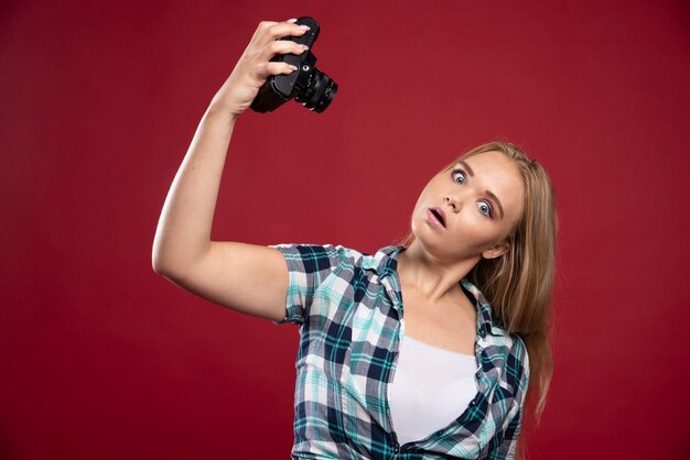 Giovane fotografia bionda che tiene una macchina fotografica professionale e prende il suo selfie in posizioni strane.