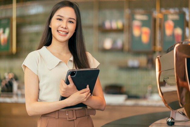 Giovane freelance creativo digitale nomade asiatico femmina sorridente ritratto in piedi mano tenere tablet dispositivo sorridente casual relax lavoro lavorando ovunque nuovo stile di vita lavorando dal ristorante caffetteria