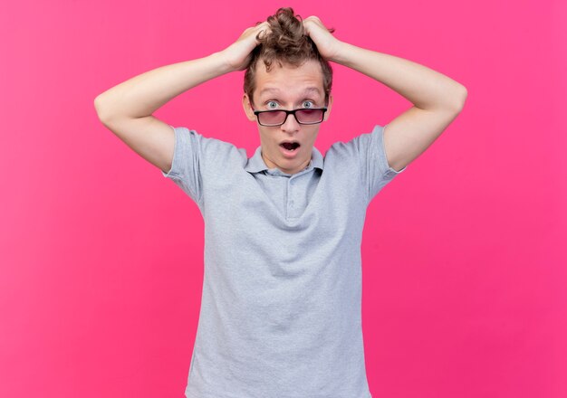 Giovane frustrato con gli occhiali neri che indossa una polo grigia che tocca la testa tirando i suoi capelli in piedi sul muro rosa